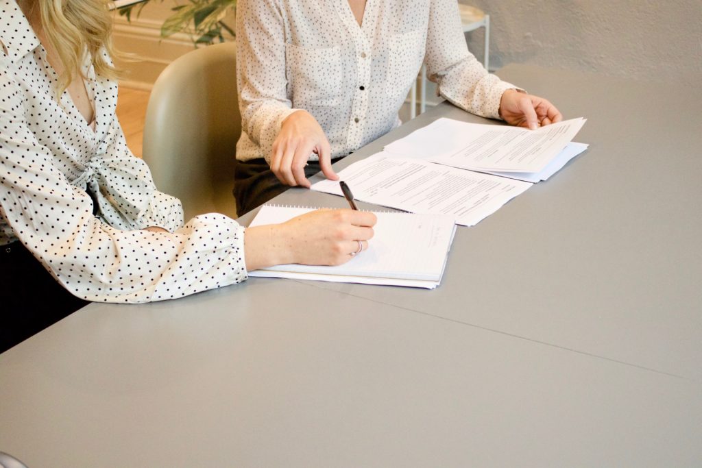 Two businesswomen sitting at a table, looking and papers and writing down notes